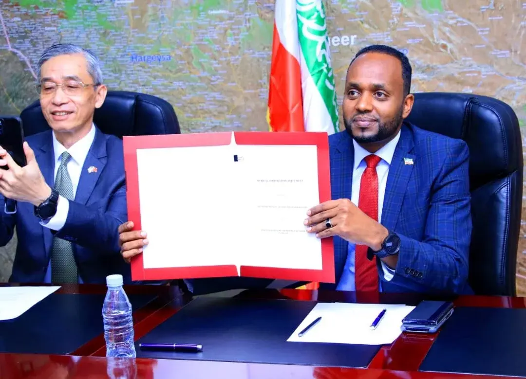 Two officials seated at a table, one holding up a signed medical cooperation agreement in a red folder